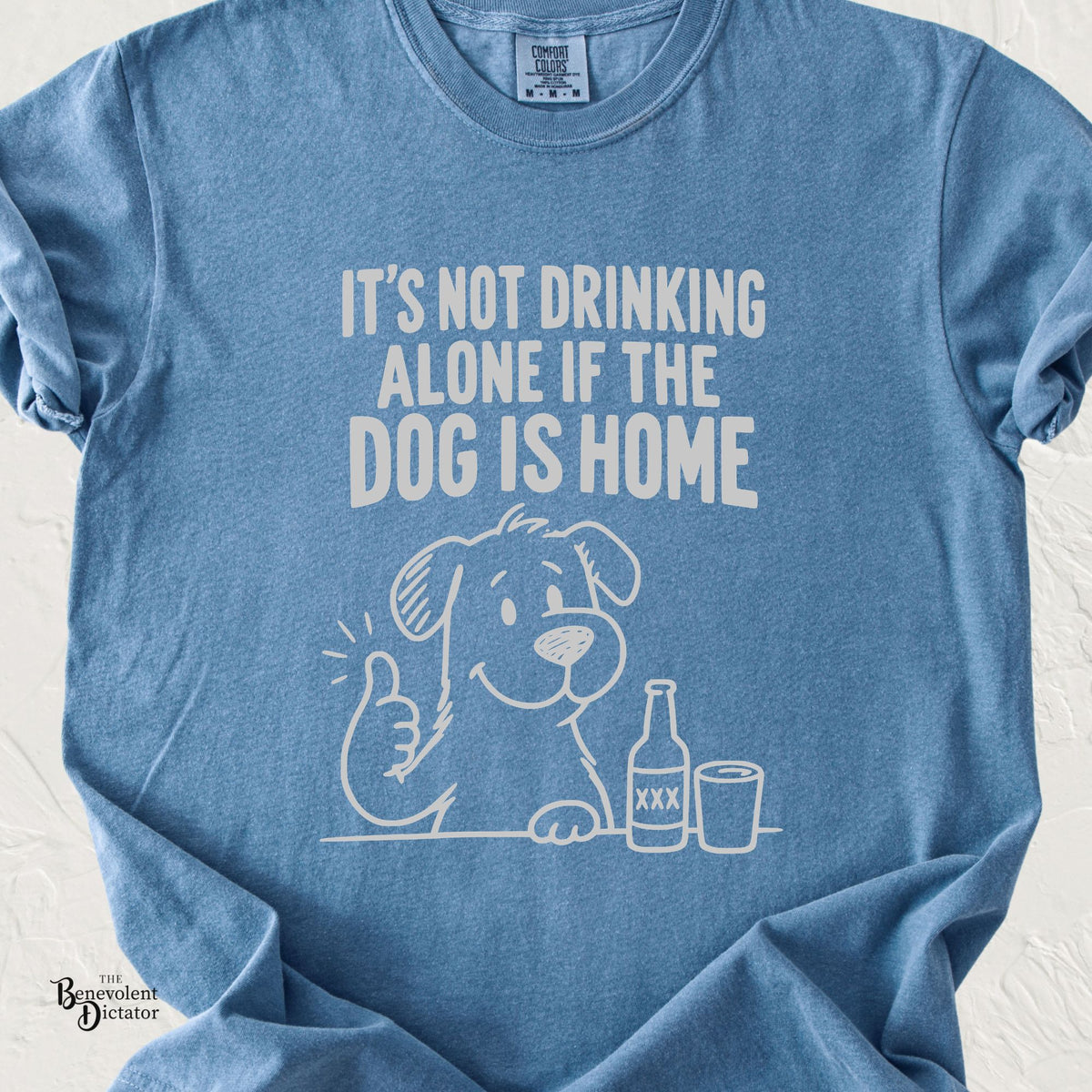A light blue crewneck t-shirt with a white graphic print that reads 'IT'S NOT DRINKING ALONE IF THE DOG IS HOME' above a cartoon dog giving a thumbs-up next to a beer bottle and a glass. The shirt is laid flat on a white textured background.