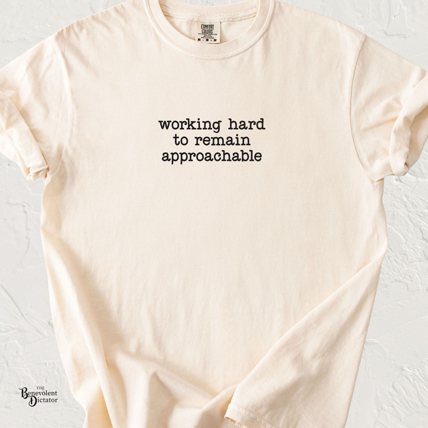 Ivory Comfort Colors t-shirt featuring minimalist black typewriter text that reads “working hard to remain approachable.”