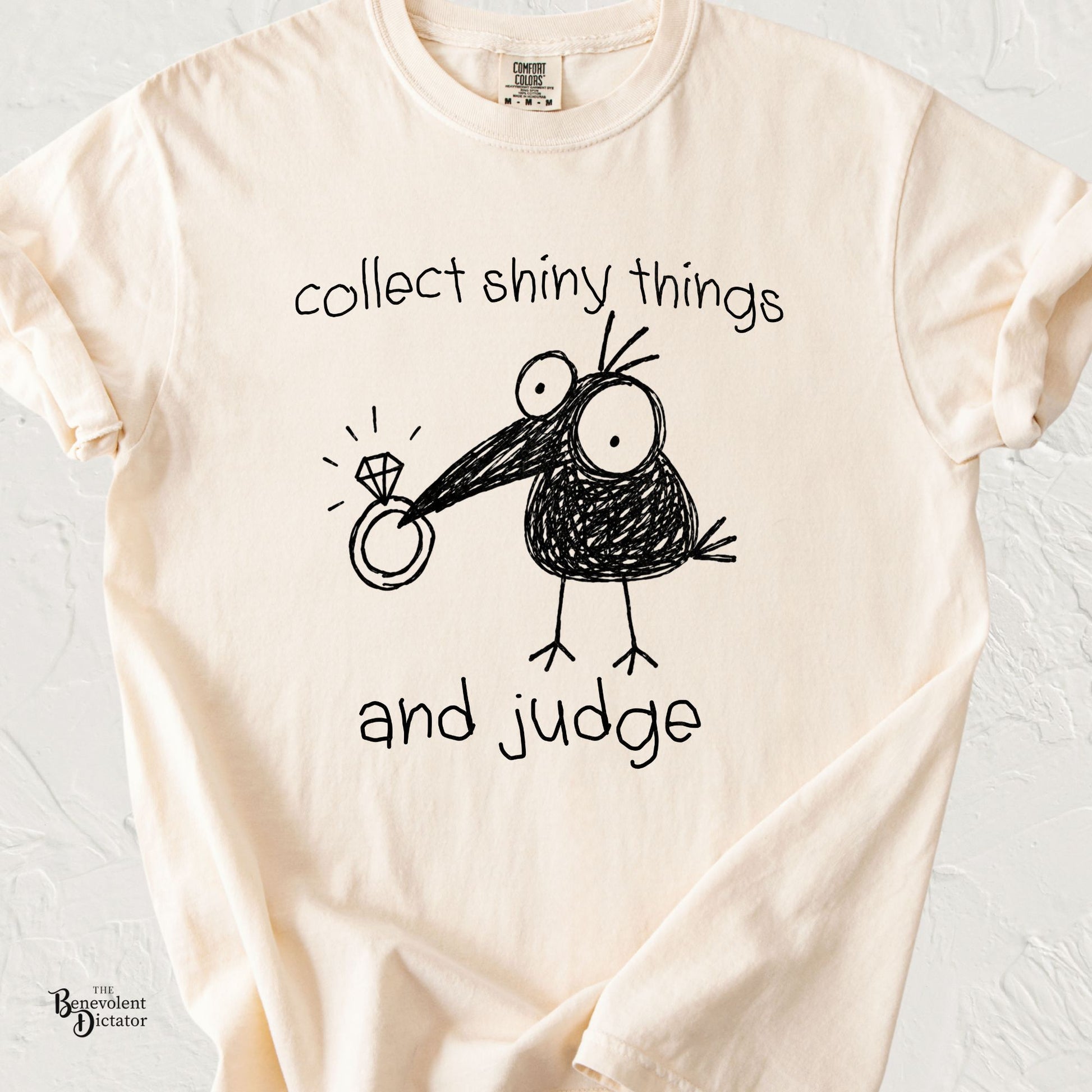 Ivory Comfort Colors t-shirt featuring a quirky doodle crow holding a shiny ring with the phrase “collect shiny things and judge” in playful handwritten text.