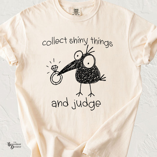 Ivory Comfort Colors t-shirt featuring a quirky doodle crow holding a shiny ring with the phrase “collect shiny things and judge” in playful handwritten text.