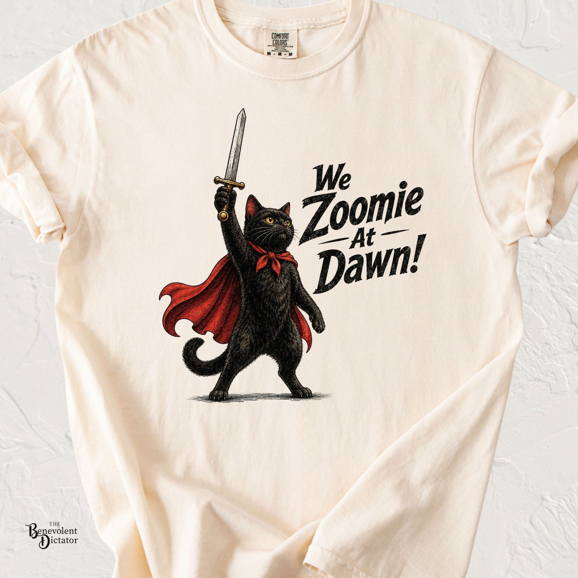 Vintage-inspired illustration of a black cat wearing a flowing red cape and raising a sword triumphantly beside the words "We Zoomie at Dawn!" on an ivory Comfort Colors T-shirt.