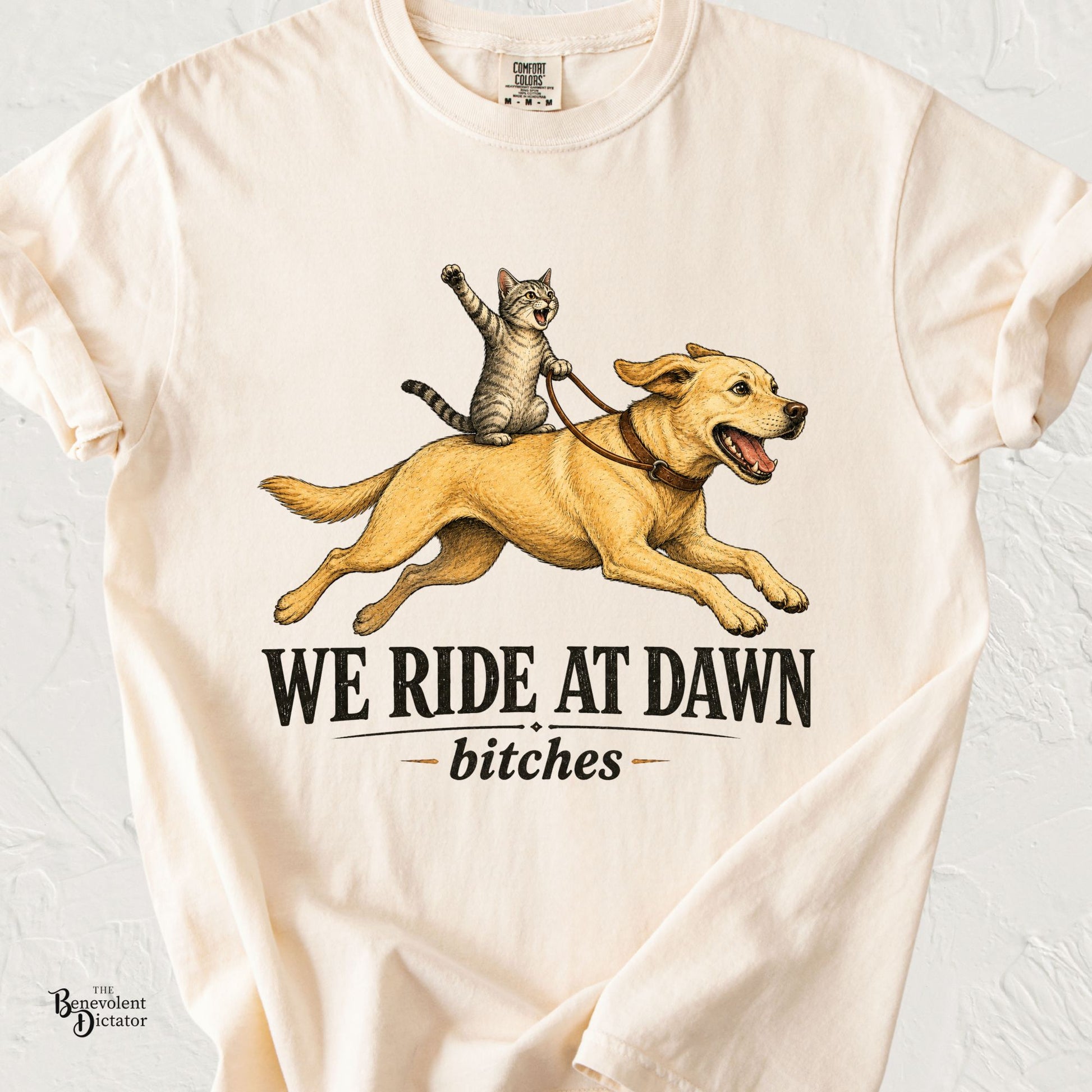 Ivory Comfort Colors t-shirt featuring a cat riding a running Labrador with one paw raised above the phrase "We Ride at Dawn Bitches" in vintage lettering.