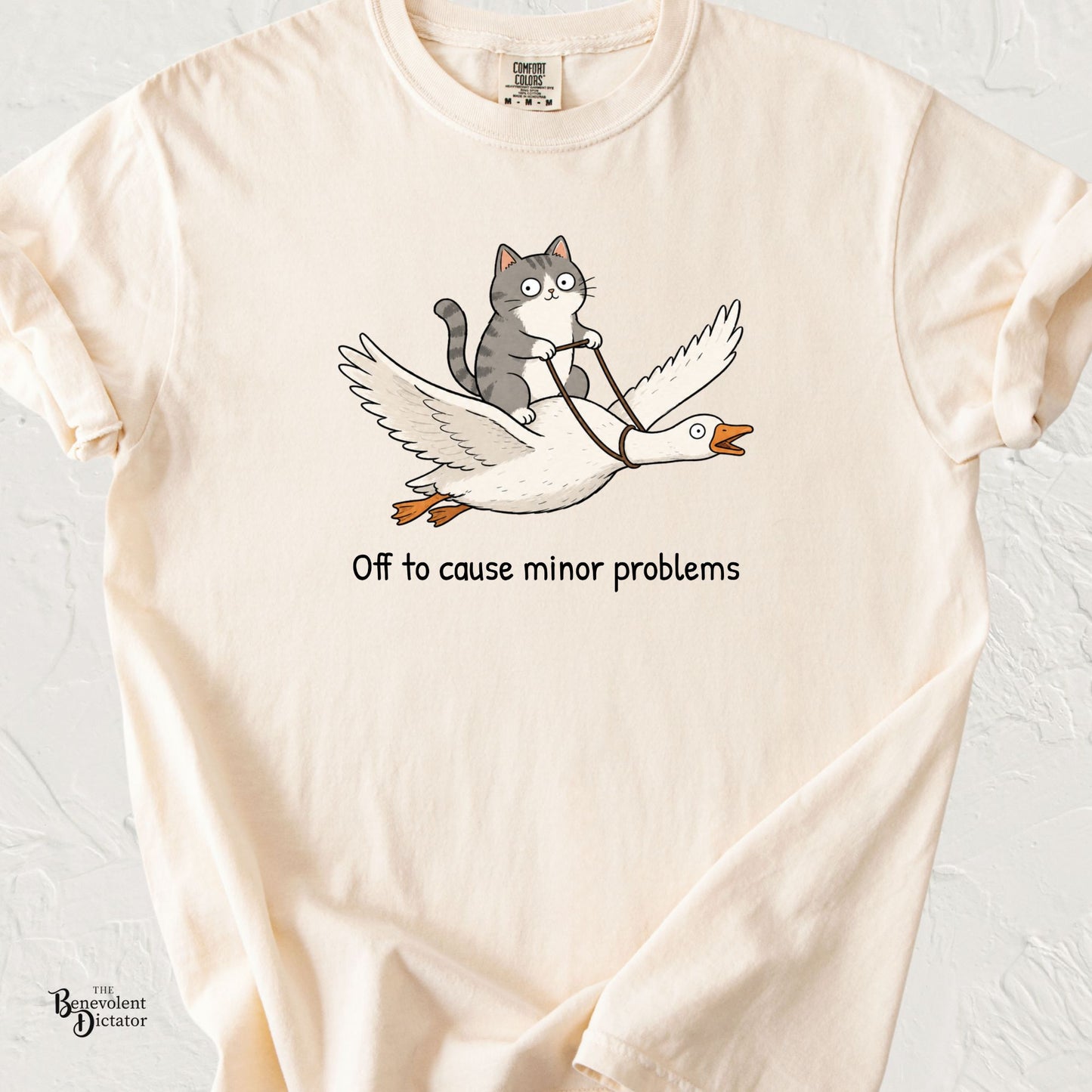 Cute illustration of a wide-eyed gray cat riding a flying goose with the phrase "Off to Cause Minor Problems" on a Comfort Colors® t-shirt.
