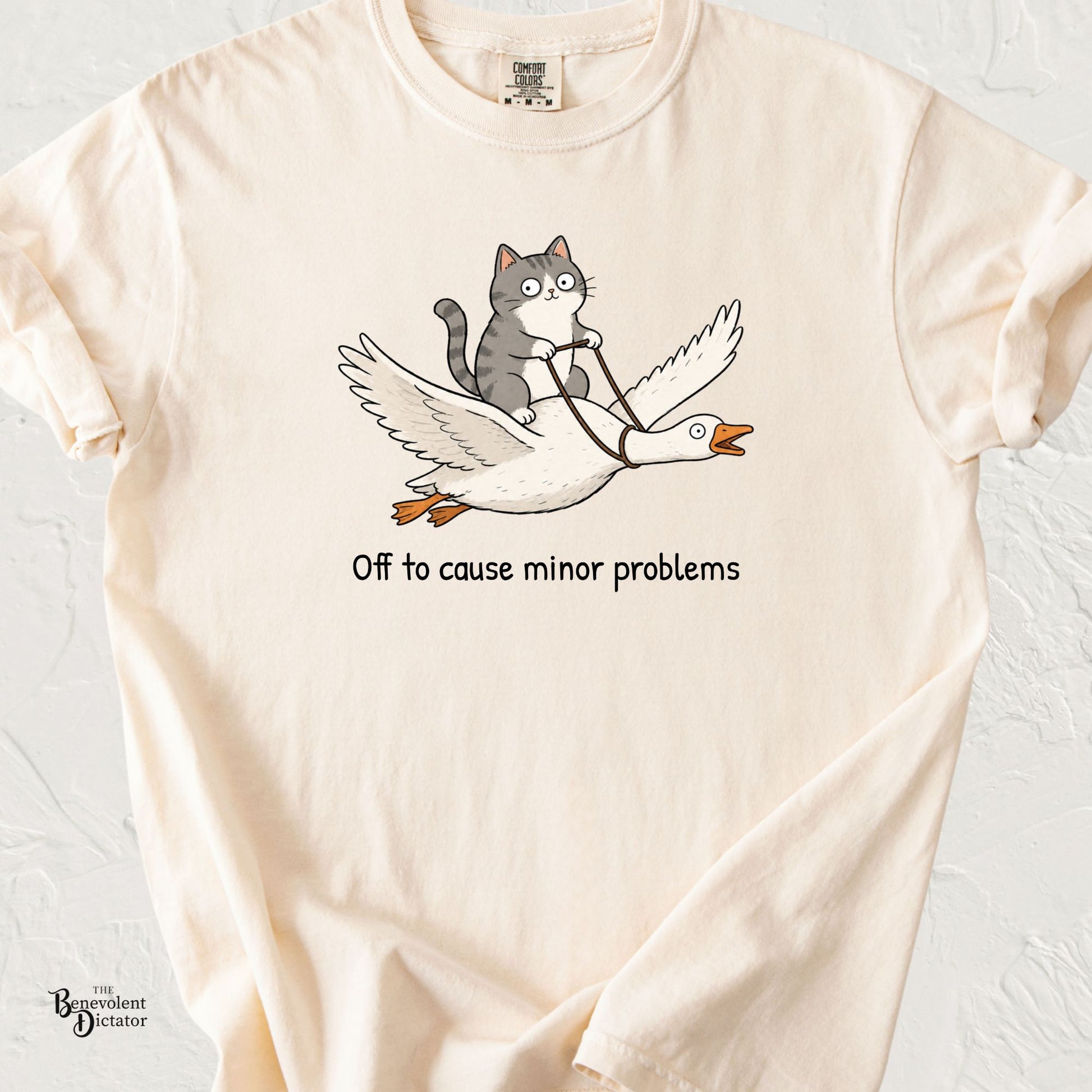 Cute illustration of a wide-eyed gray cat riding a flying goose with the phrase "Off to Cause Minor Problems" on a Comfort Colors® t-shirt.