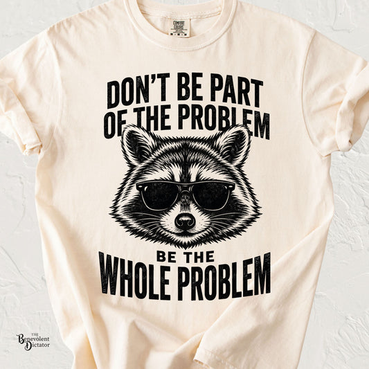 A cream-colored short-sleeve t-shirt with a black graphic print of a raccoon wearing sunglasses, surrounded by the text 'DON'T BE PART OF THE PROBLEM BE THE WHOLE PROBLEM'.