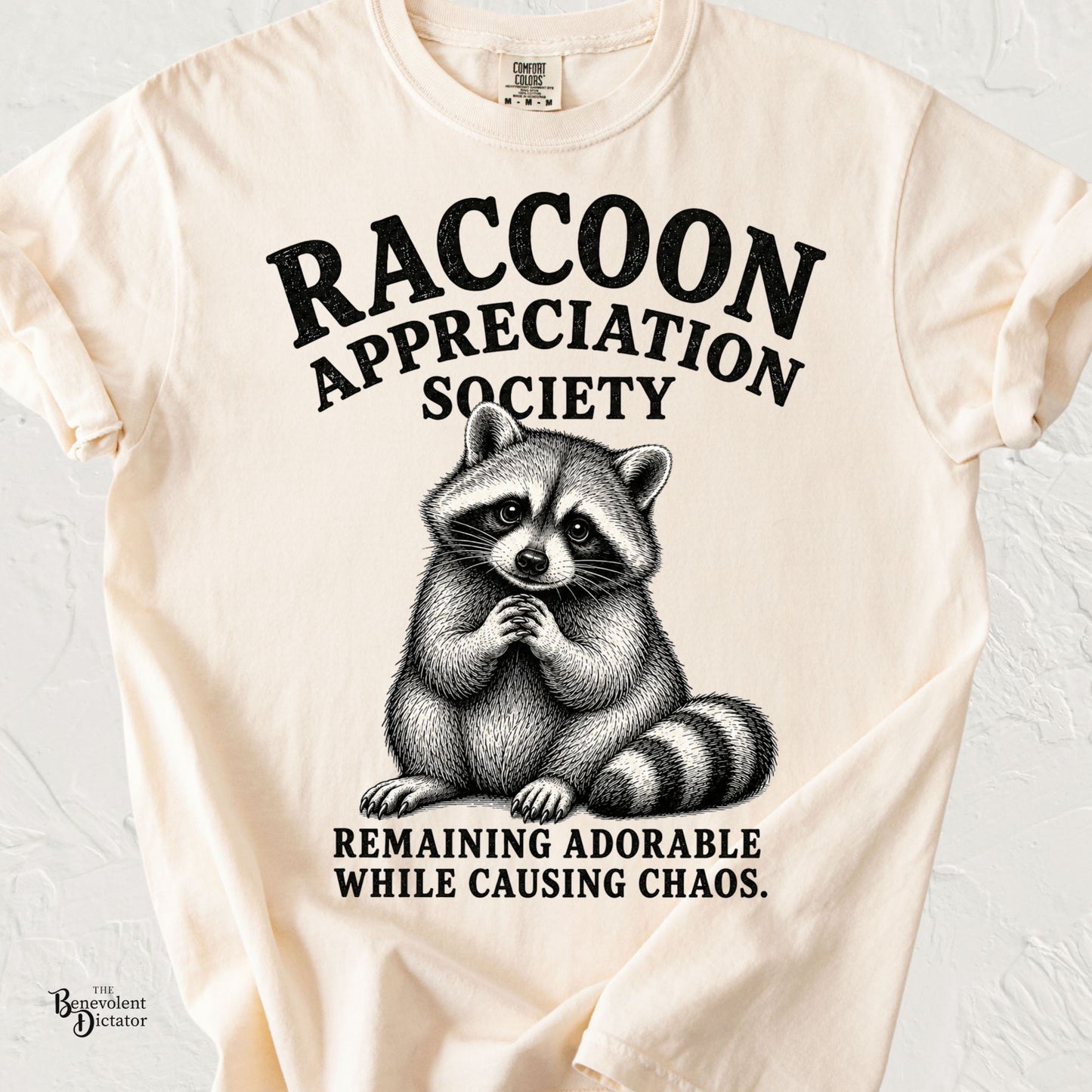 Vintage-inspired raccoon illustration with clasped paws and the text "Raccoon Appreciation Society" and "Remaining Adorable While Causing Chaos."