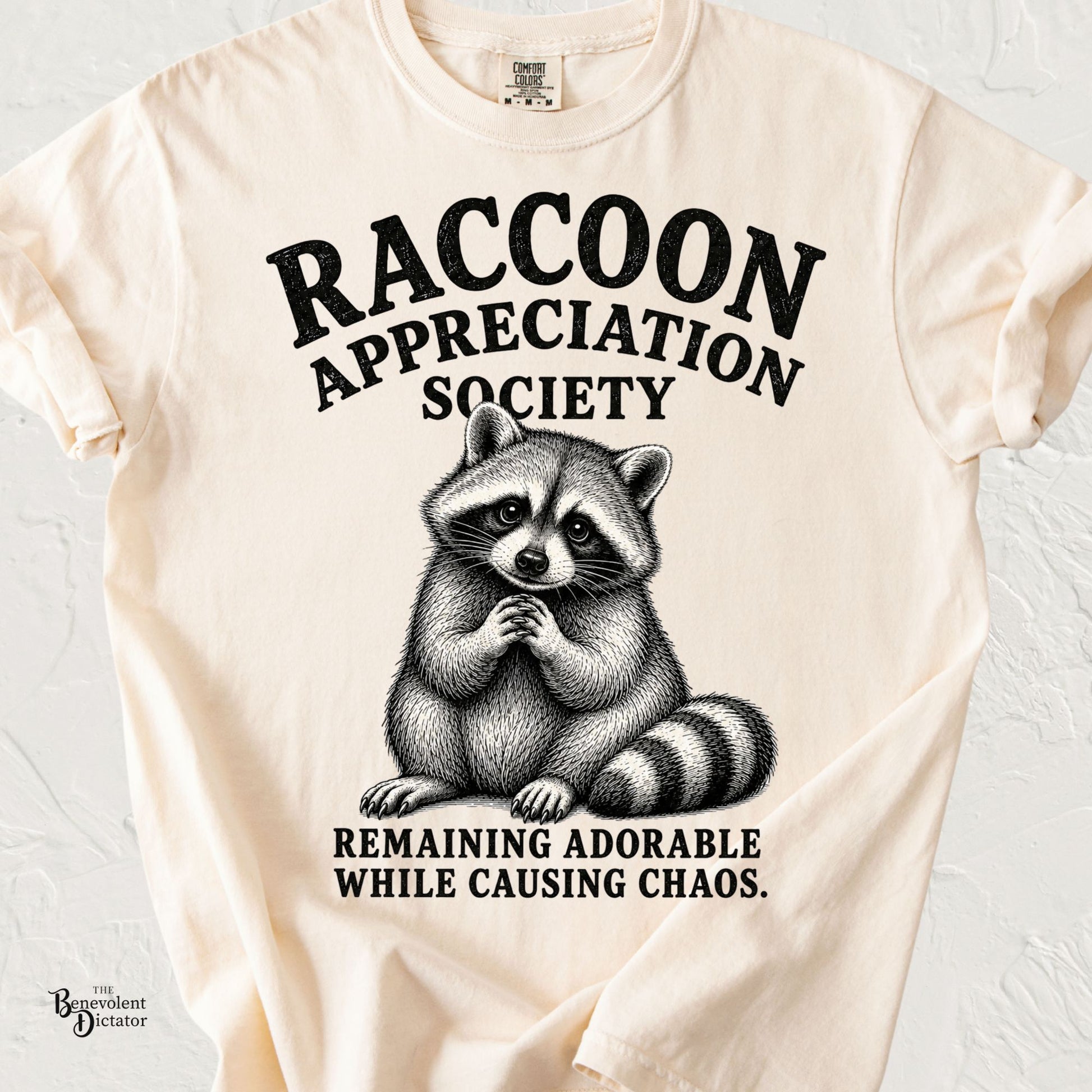 Vintage-inspired raccoon illustration with clasped paws and the text "Raccoon Appreciation Society" and "Remaining Adorable While Causing Chaos."