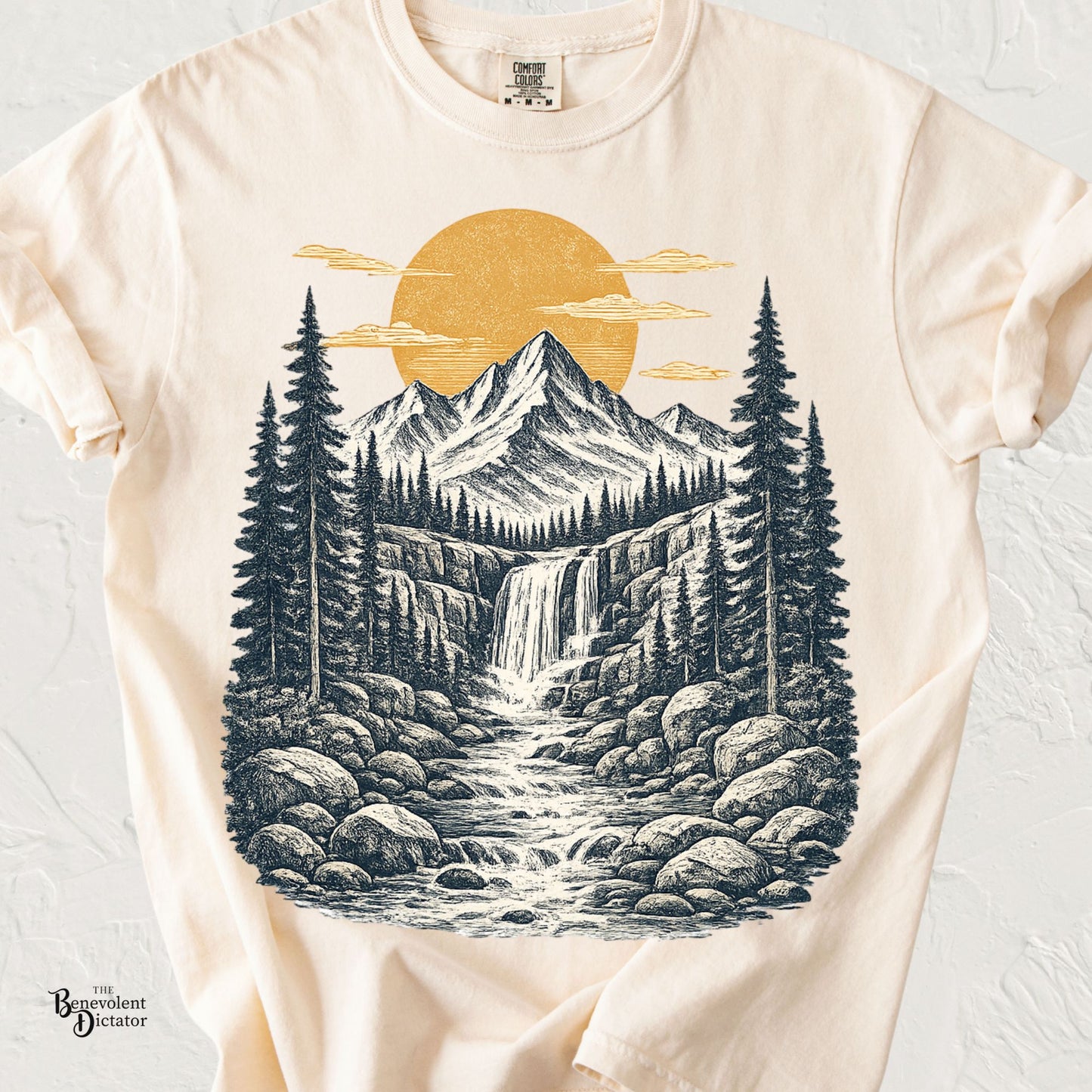 Vintage mountain waterfall illustration with pine trees, flowing stream, and golden sun printed on a Comfort Colors nature shirt.