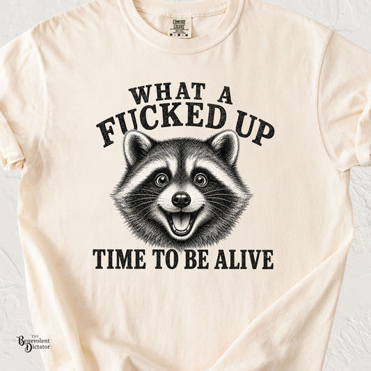 Ivory Comfort Colors t-shirt featuring an excited vintage engraved raccoon with the phrase “What a Fucked Up Time to Be Alive” in distressed black typography.