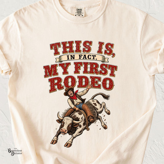 An off-white short-sleeve t-shirt with a vintage-style western graphic that reads 'THIS IS, IN FACT. MY FIRST RODEO' above an illustration of a cowboy riding a bucking bull.