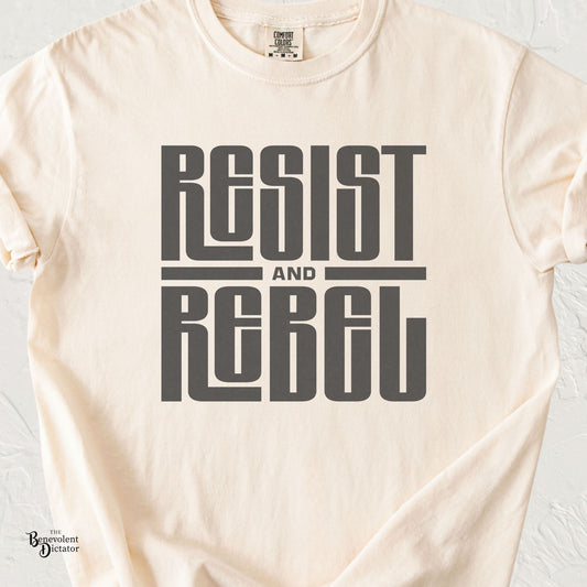 A cream-colored short-sleeve t-shirt with a dark gray graphic print reading 'RESIST AND REBEL' on the front, laid flat against a light textured background.