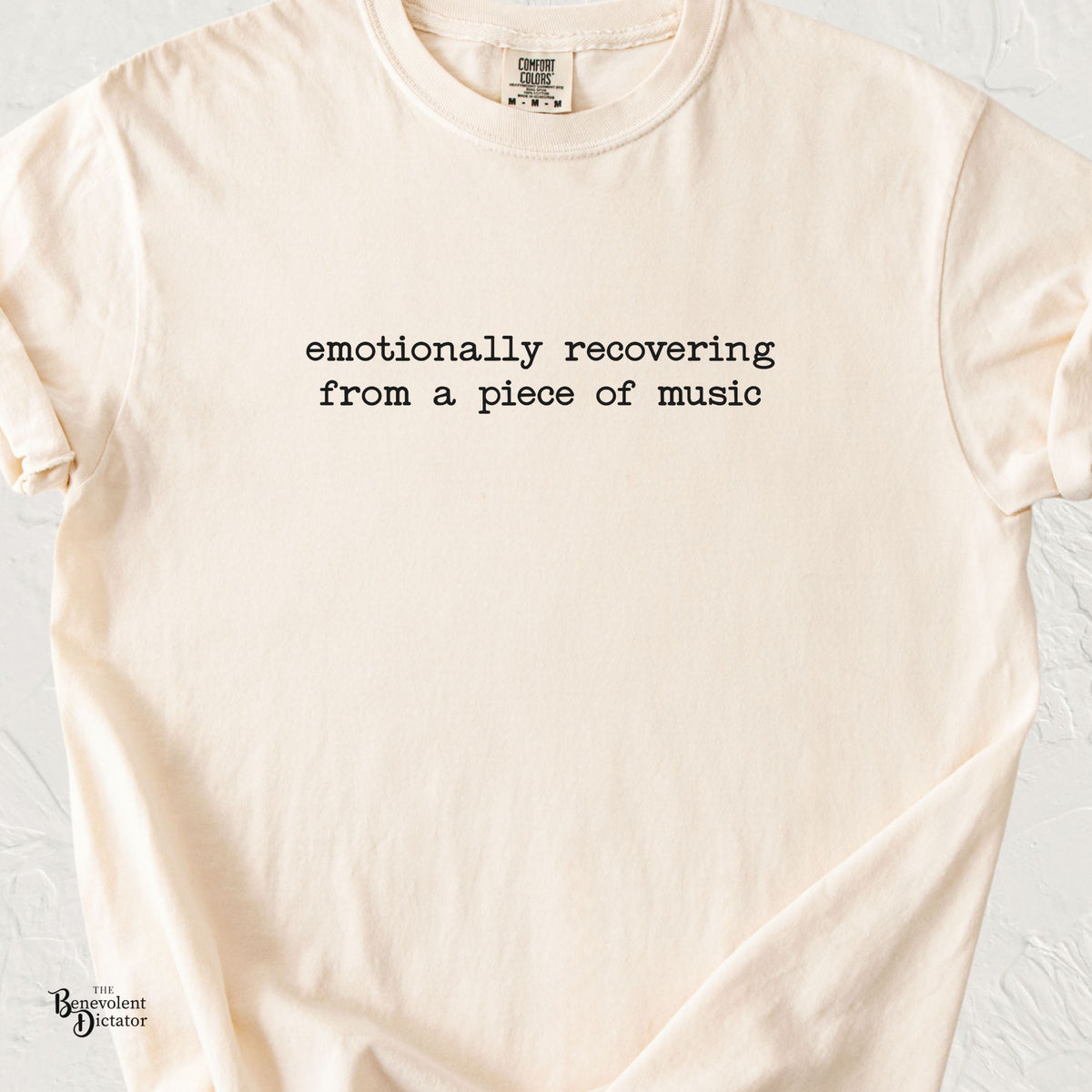 A cream-colored short-sleeve t-shirt with the humorous text 'emotionally recovering from a piece of music' printed in black typewriter font across the chest. The shirt is laid flat on a light, textured background.