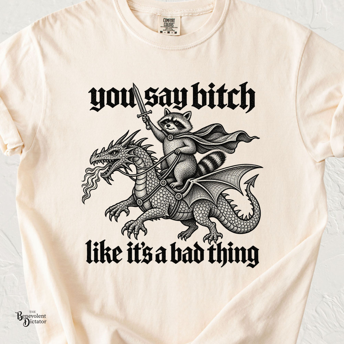 Ivory Comfort Colors t-shirt featuring a sword-wielding raccoon riding a fire-breathing dragon with the phrase “You Say Bitch Like It’s a Bad Thing” in bold medieval lettering.