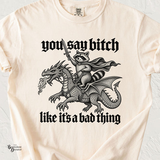 Ivory Comfort Colors t-shirt featuring a sword-wielding raccoon riding a fire-breathing dragon with the phrase “You Say Bitch Like It’s a Bad Thing” in bold medieval lettering.