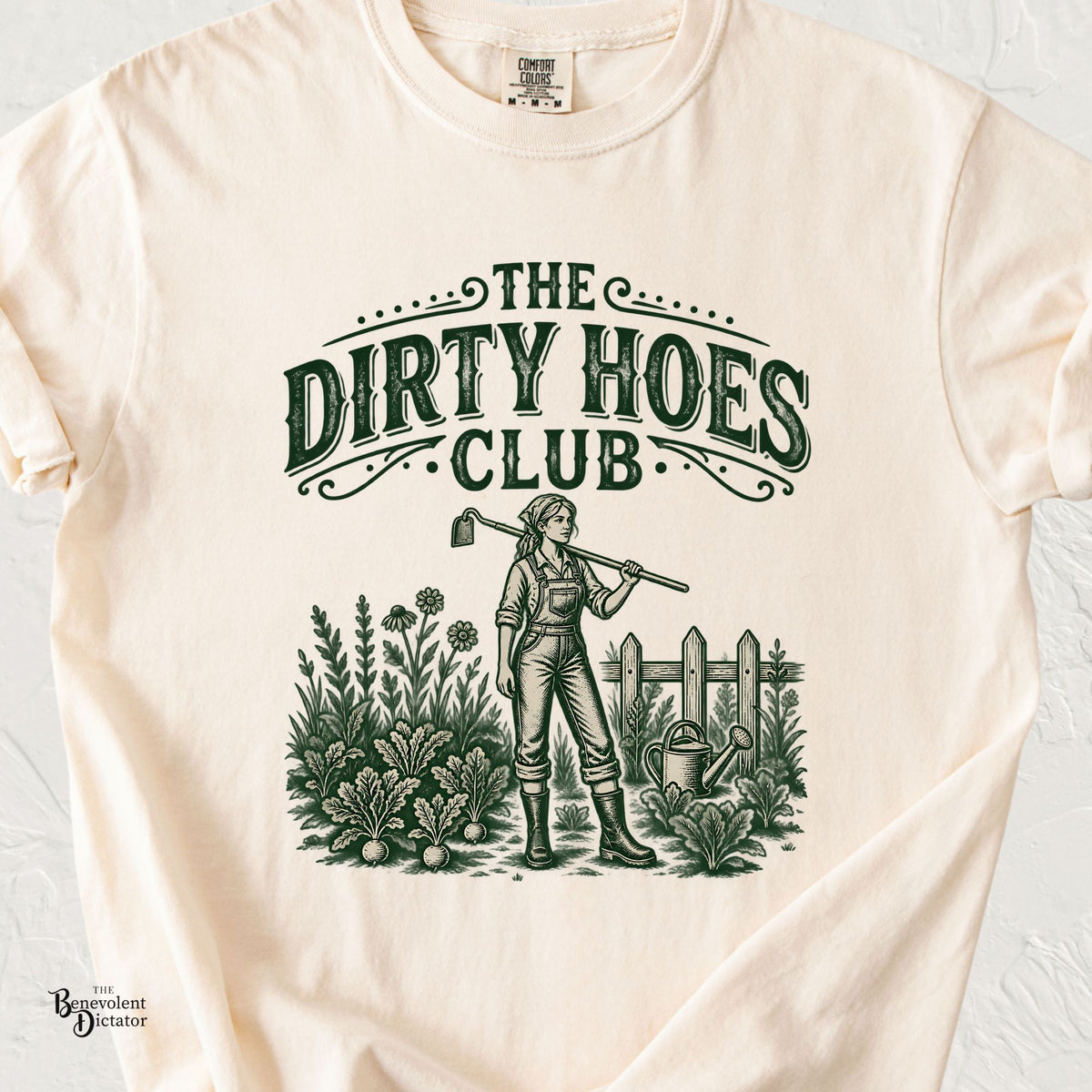 An ivory Comfort Colors 1717 short-sleeve t-shirt with a vintage-style green graphic that reads 'THE DIRTY HOES CLUB' above an illustration of a woman in overalls gardening with a hoe, watering can, and fence.