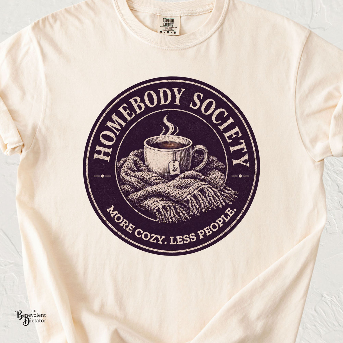An off-white crewneck t-shirt featuring a circular graphic print with a coffee cup wrapped in a blanket and the text 'HOMEBODY SOCIETY' and 'MORE COZY. LESS PEOPLE.