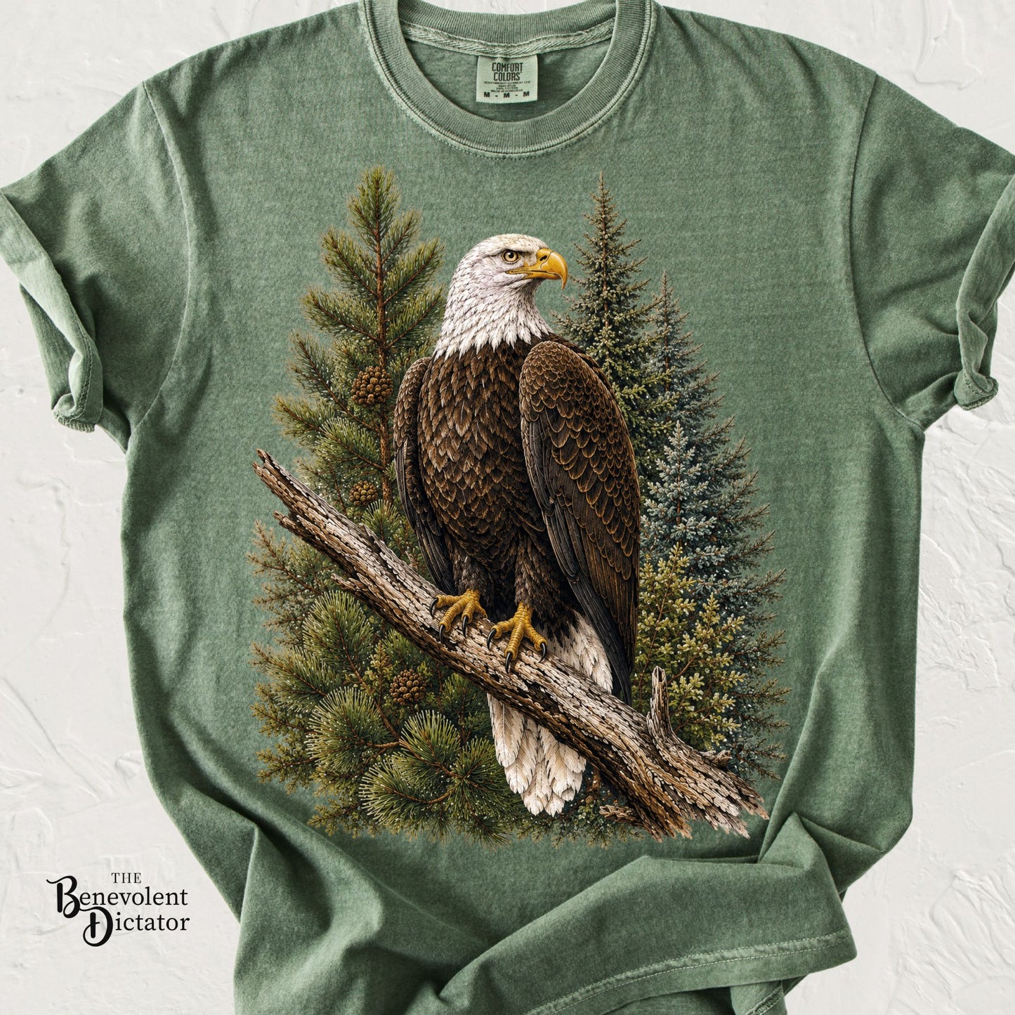 A sage green heathered t-shirt featuring a large graphic print of a bald eagle perched on a tree branch in front of pine trees. 