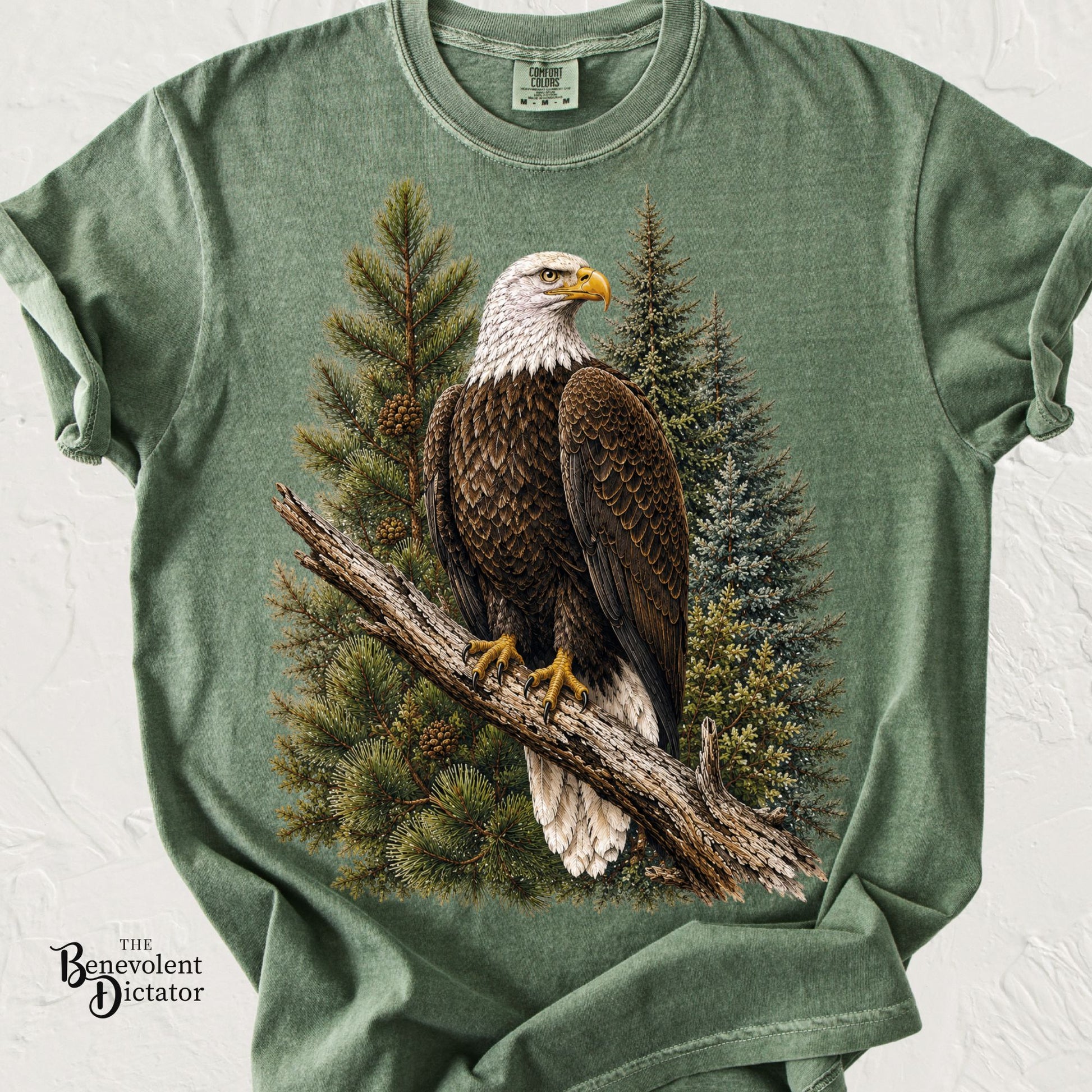 A sage green heathered t-shirt featuring a large graphic print of a bald eagle perched on a tree branch in front of pine trees. 