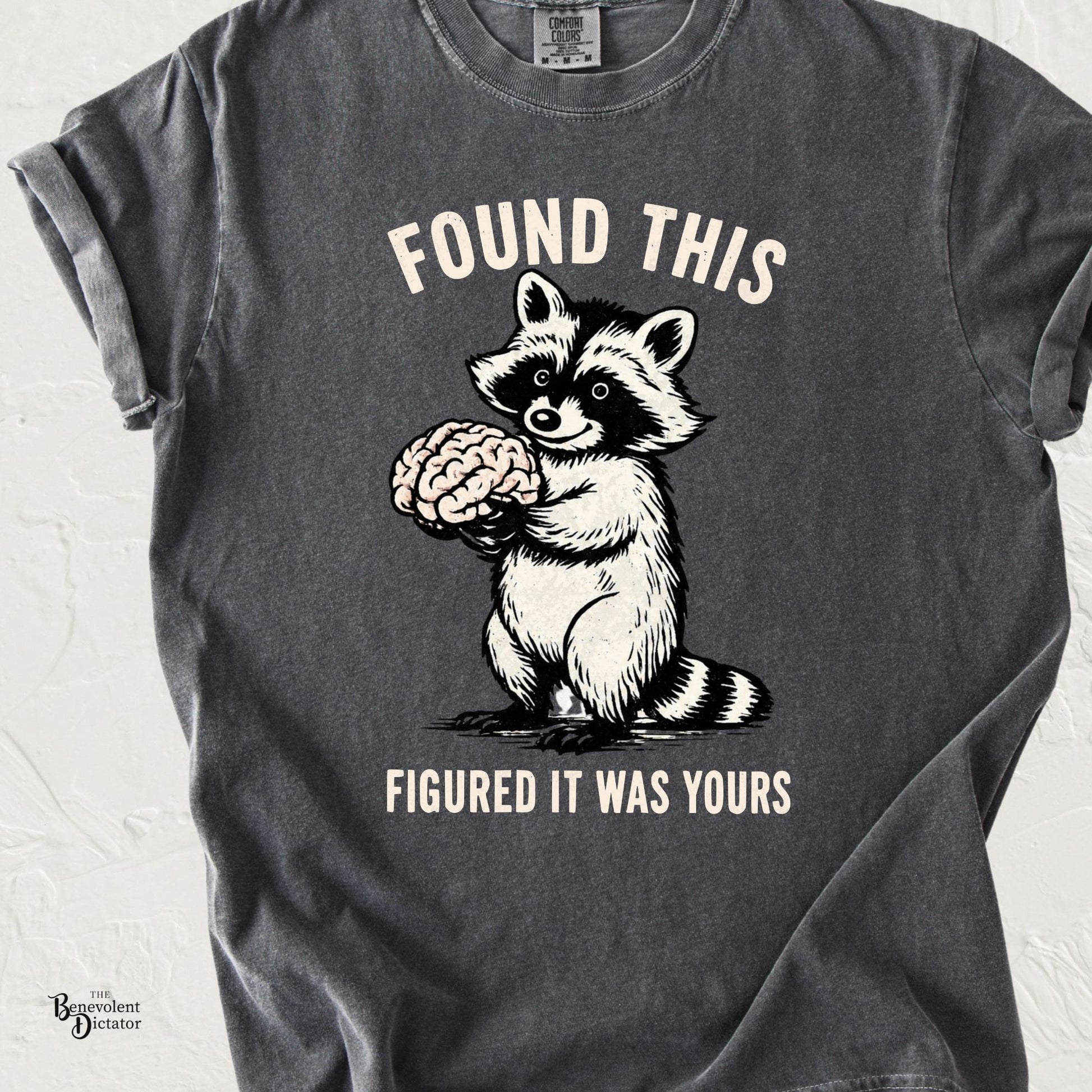 Funny raccoon graphic t-shirt featuring a raccoon holding a brain with the text “Found This Figured It Was Yours” on a pepper gray shirt.