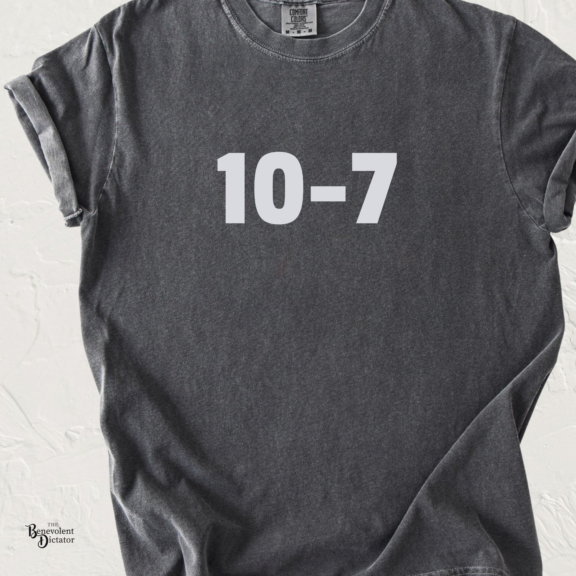 Dark gray graphic t-shirt featuring light grey “10-7” text centered on the chest, shown in a lifestyle mockup.