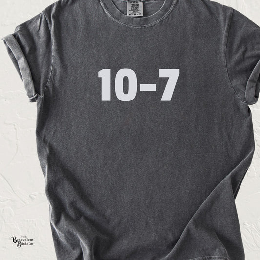 Dark gray graphic t-shirt featuring light grey “10-7” text centered on the chest, shown in a lifestyle mockup.