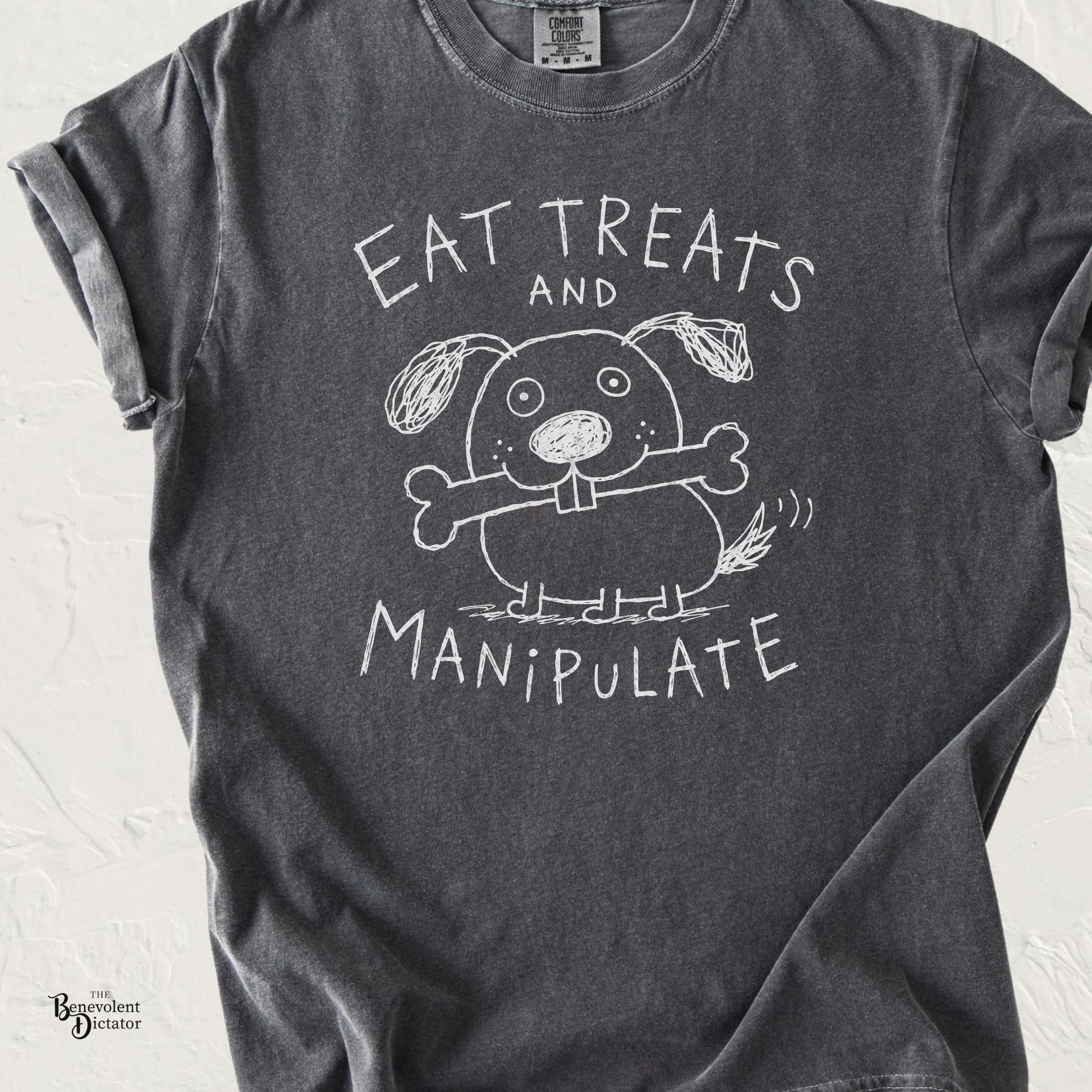 Pepper gray graphic t-shirt featuring a childlike doodle dog holding a bone with the phrase “Eat Treats and Manipulate” in hand-drawn lettering.