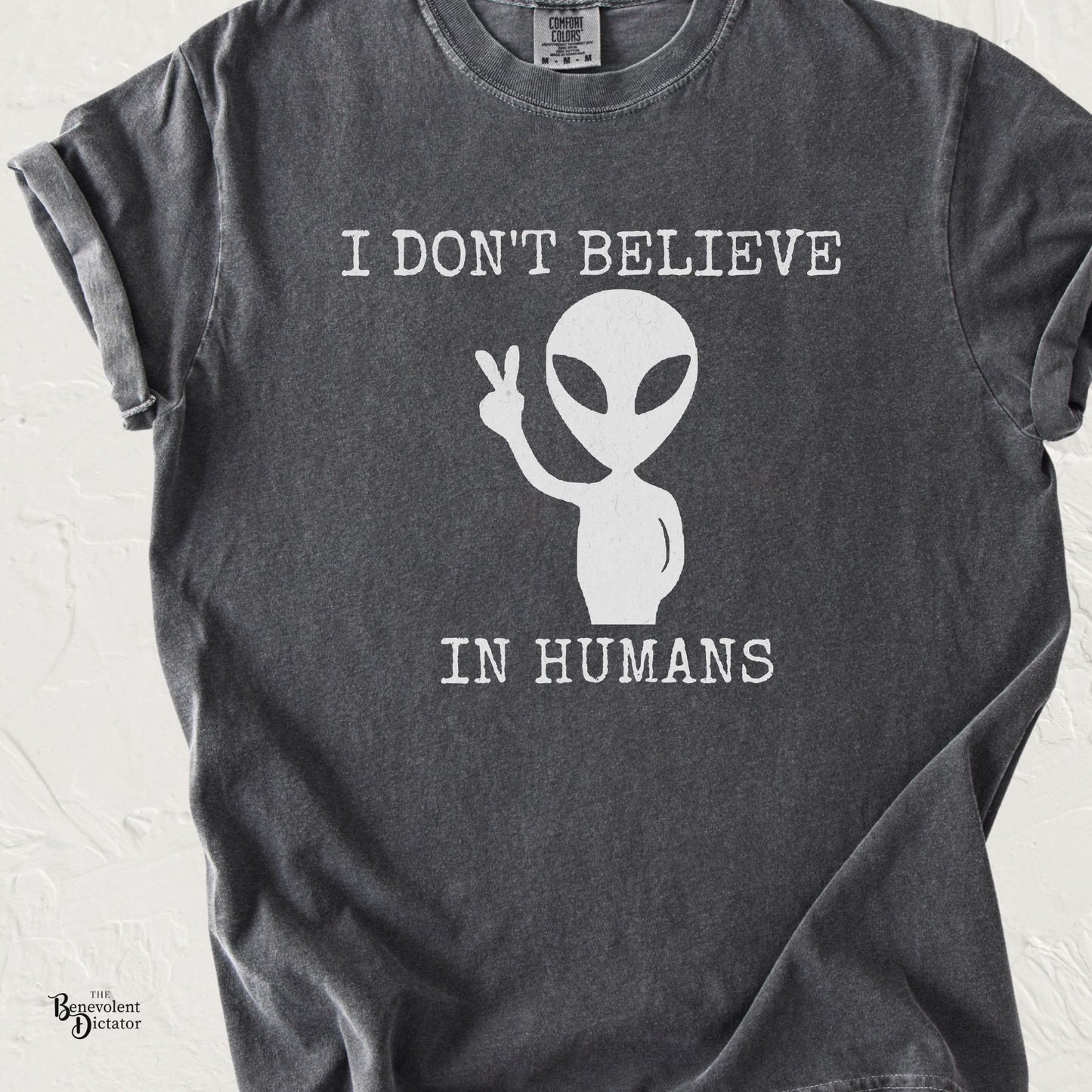 Pepper gray Comfort Colors t-shirt featuring a minimalist alien graphic flashing a peace sign with the phrase “I Don’t Believe in Humans.”