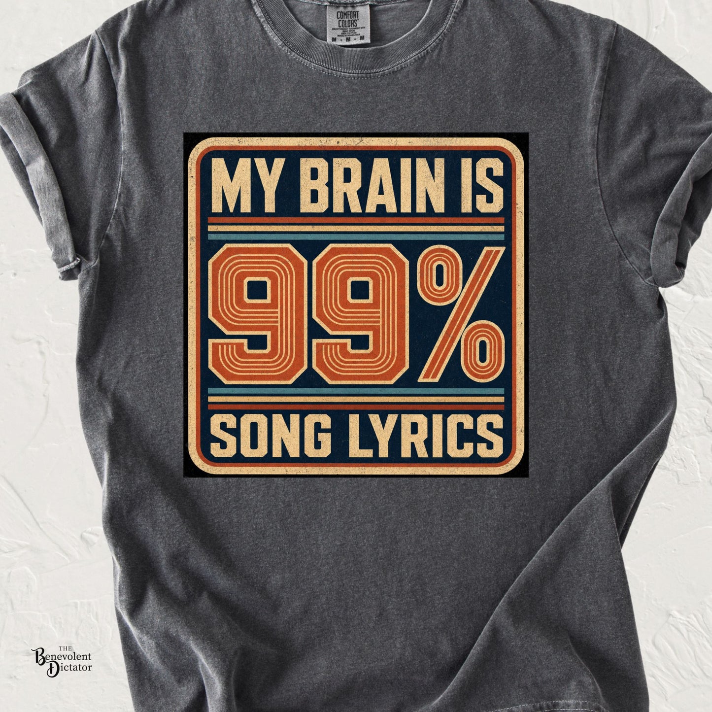 Pepper Comfort Colors t-shirt featuring a retro distressed badge graphic in cream, navy, orange, and muted blue with bold text reading “My Brain Is 99% Song Lyrics.”