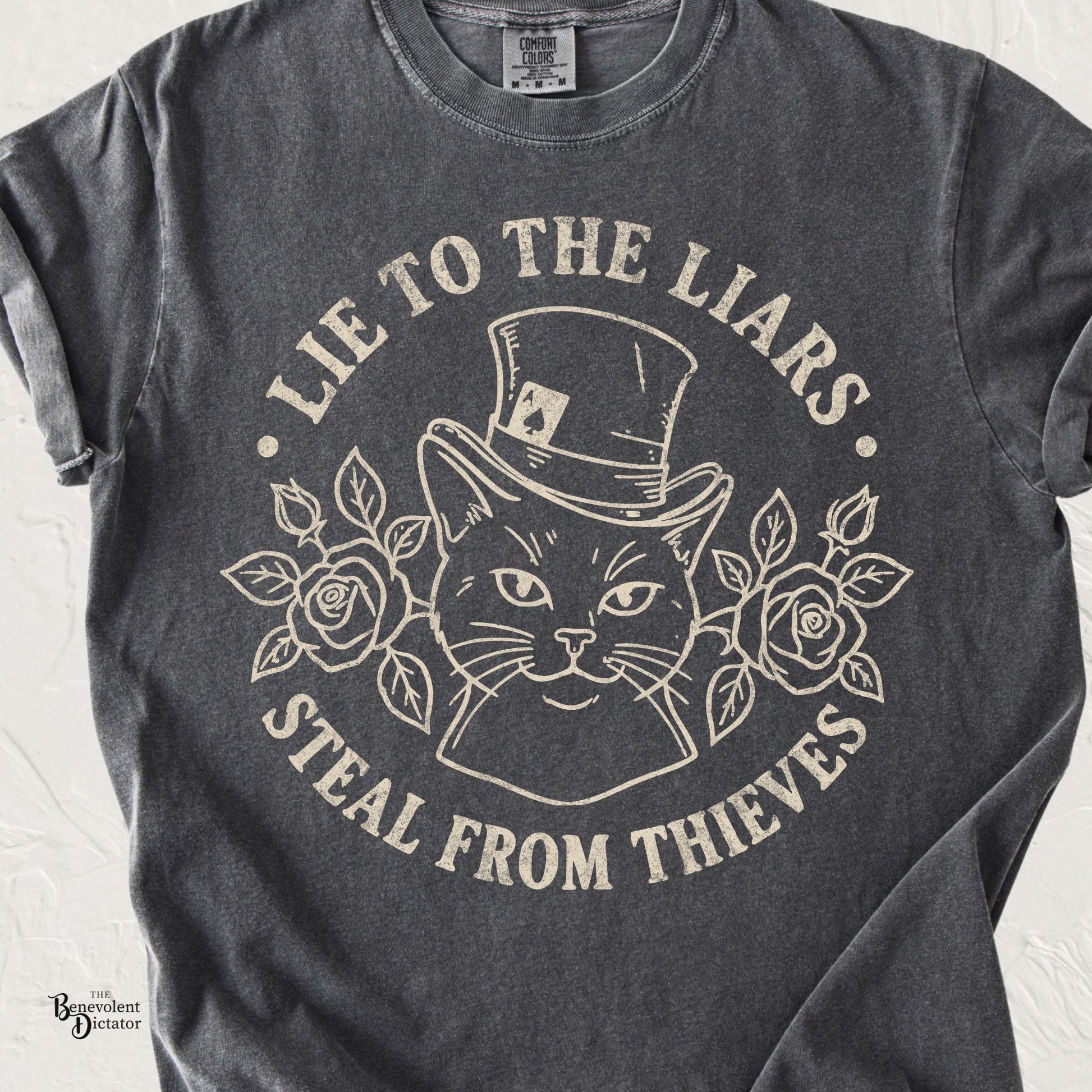 Vintage cream line art of a sly cat wearing a top hat surrounded by roses with the phrase Lie to the Liars, Steal from Thieves on a pepper Comfort Colors t-shirt.