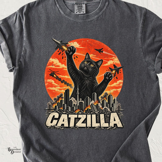 Pepper Comfort Colors t-shirt featuring a giant black cat swatting fighter jets above a burning city with an orange-red sky and bold “CATZILLA” text.