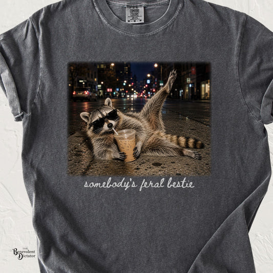 Pepper Comfort Colors t-shirt featuring a sunglasses-wearing raccoon sprawled on a city street at night drinking iced coffee, with handwritten text reading “somebody’s feral bestie.”