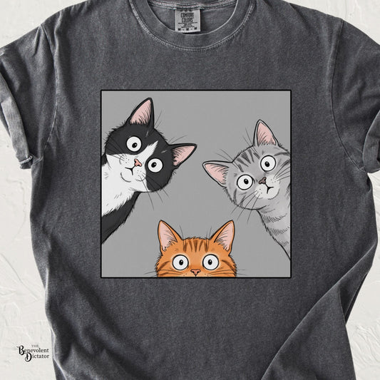 Pepper Comfort Colors t-shirt featuring three wide-eyed cats peeking into a square frame, including a tuxedo cat, grey tabby, and orange tabby.