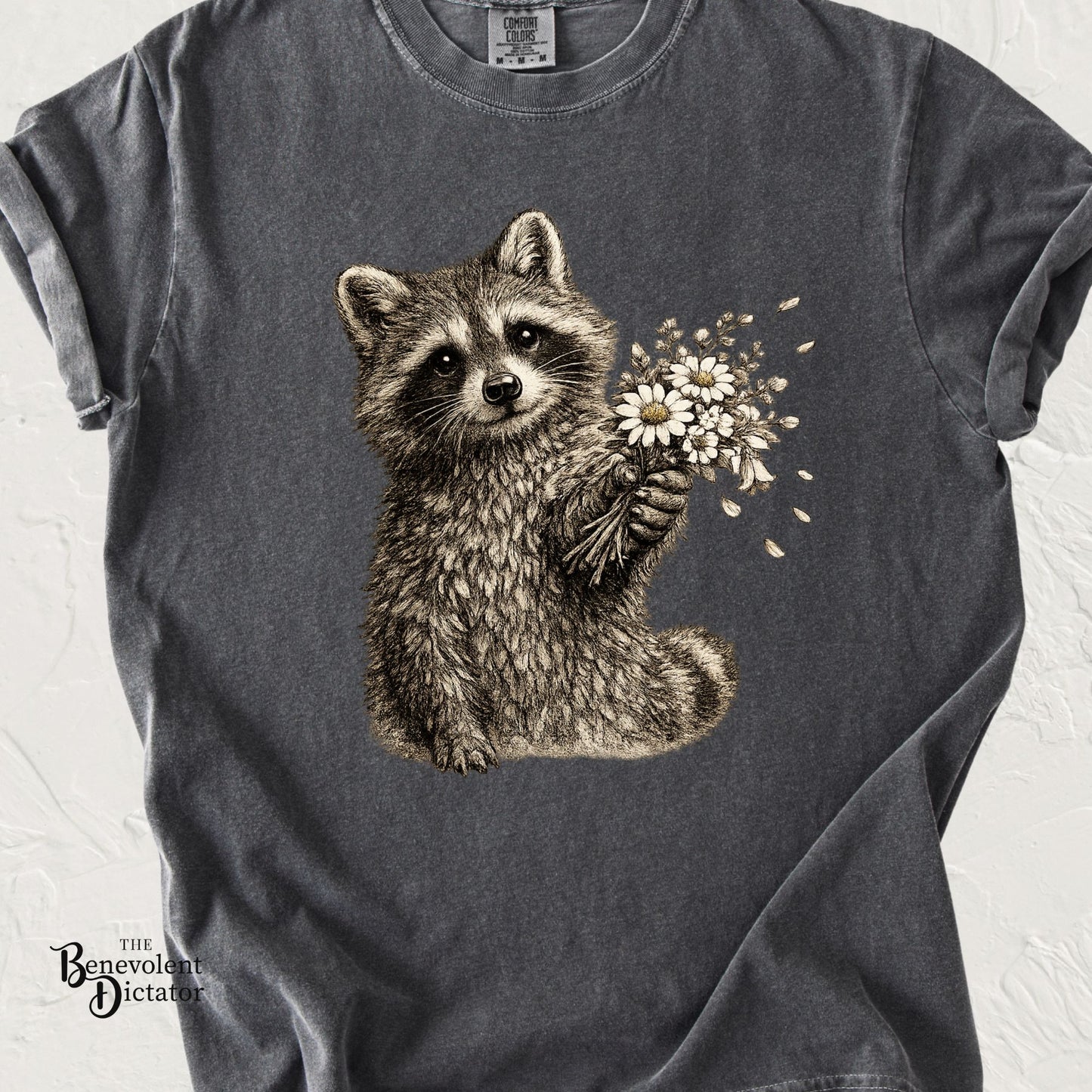Vintage-style t-shirt featuring a raccoon holding a bouquet of wildflowers in detailed woodland artwork.