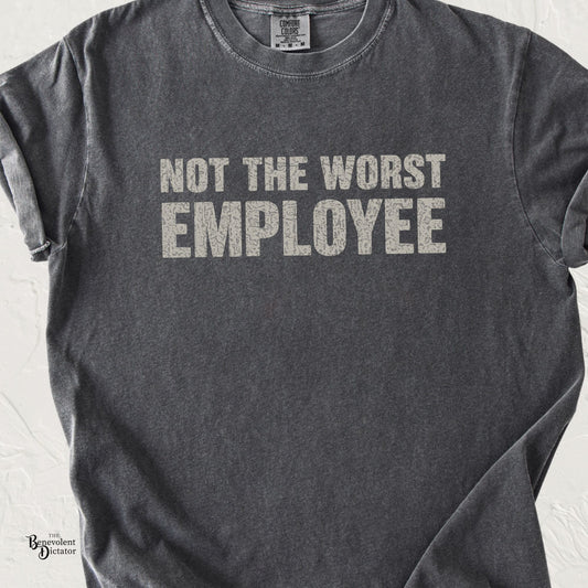 A dark gray heathered t-shirt with the phrase 'NOT THE WORST EMPLOYEE' printed in distressed white capital letters across the chest, laid flat against a light textured background.