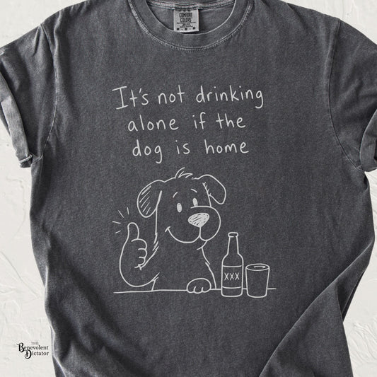 A dark gray graphic t-shirt featuring a white hand-drawn style illustration of a dog giving a thumbs-up next to a beer bottle and a glass, with the text 'It's not drinking alone if the dog is home'.