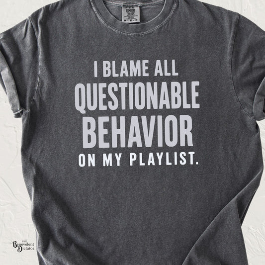 A dark gray heathered t-shirt with white text that reads 'I BLAME ALL QUESTIONABLE BEHAVIOR ON MY PLAYLIST.' laid flat against a white textured background.