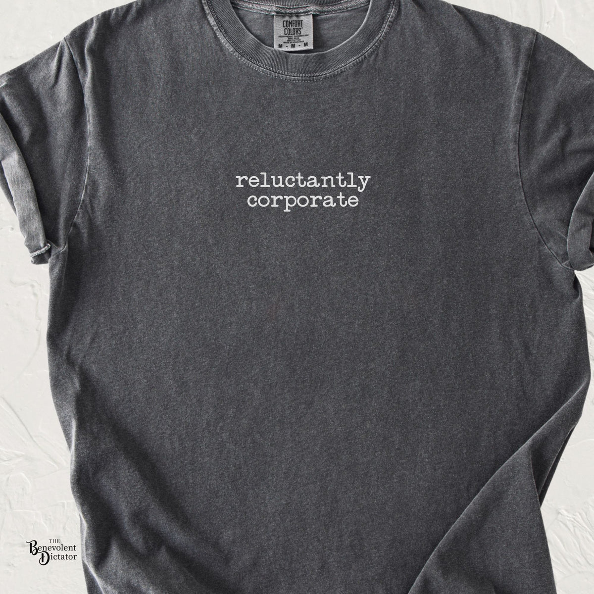 Pepper Comfort Colors t-shirt featuring the words “reluctantly corporate” in small lowercase typewriter-style lettering centered on the chest.