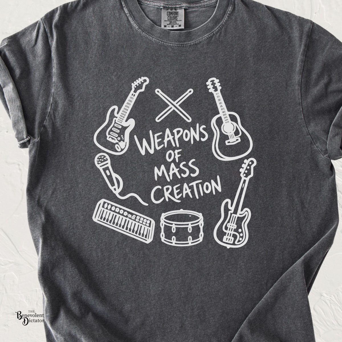 A dark gray heather t-shirt featuring a white graphic print of various musical instruments (guitars, drums, keyboard, microphone) arranged around the text 'WEAPONS OF MASS CREATION'.