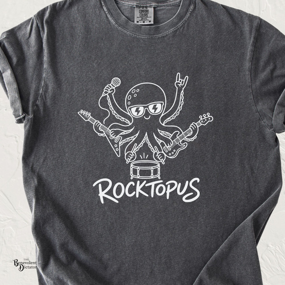 Pepper Comfort Colors t-shirt featuring a white line-art octopus wearing lightning bolt sunglasses, playing electric guitars, singing into a microphone, drumming and throwing rock horns, with the word “Rocktopus” underneath.