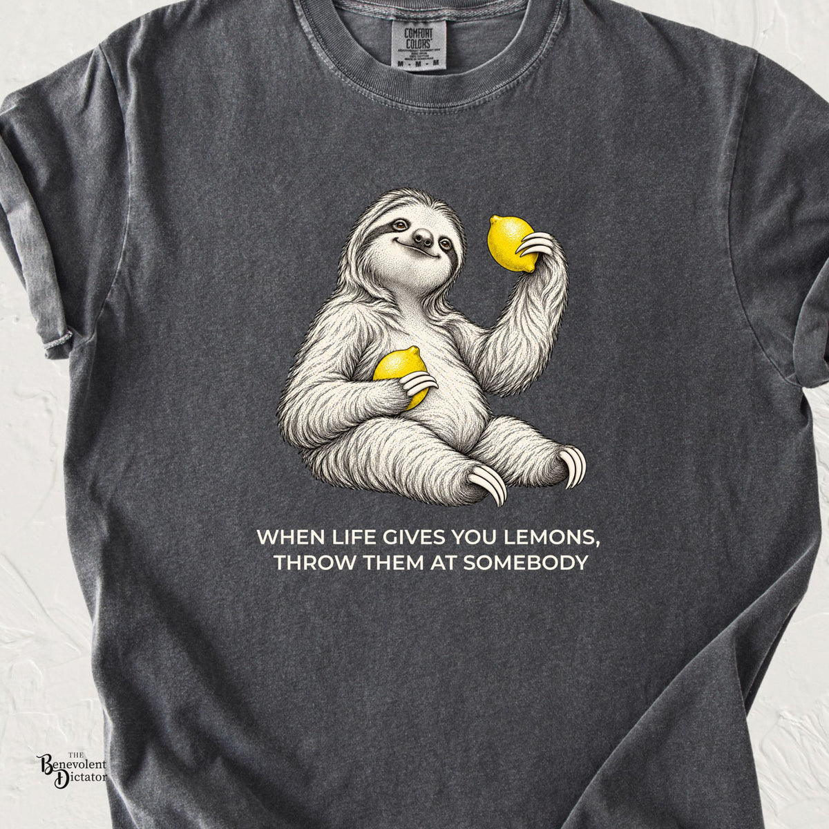 Pepper Comfort Colors t-shirt featuring a vintage engraved illustration of a smiling, seated sloth holding two bright yellow lemons. White lettering underneath reads “When Life Gives You Lemons, Throw Them at Somebody.”