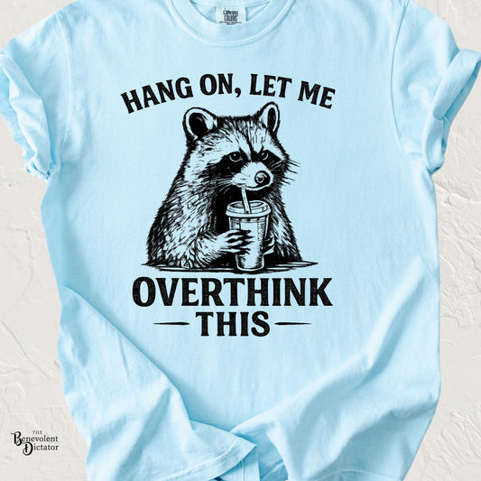 Light blue Comfort Colors t-shirt featuring a vintage-style raccoon holding a drink with the phrase "Hang On, Let Me Overthink This" in distressed black lettering.