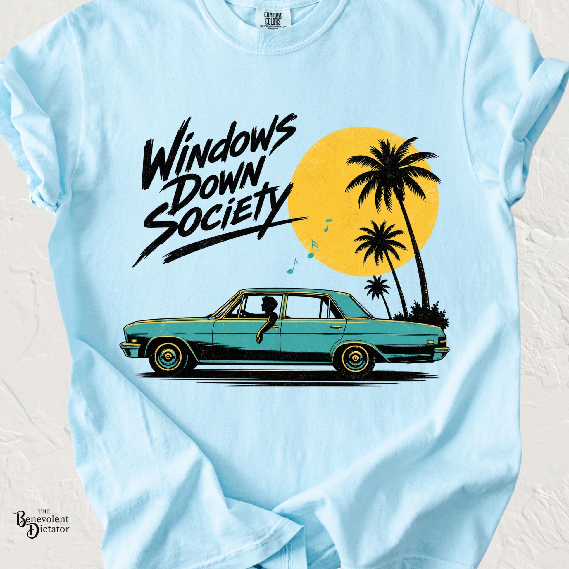 Light blue Comfort Colors t-shirt featuring a retro teal vintage sedan with a dog riding in the passenger seat, palm trees, a golden sunset, musical notes, and the phrase "Windows Down Society."