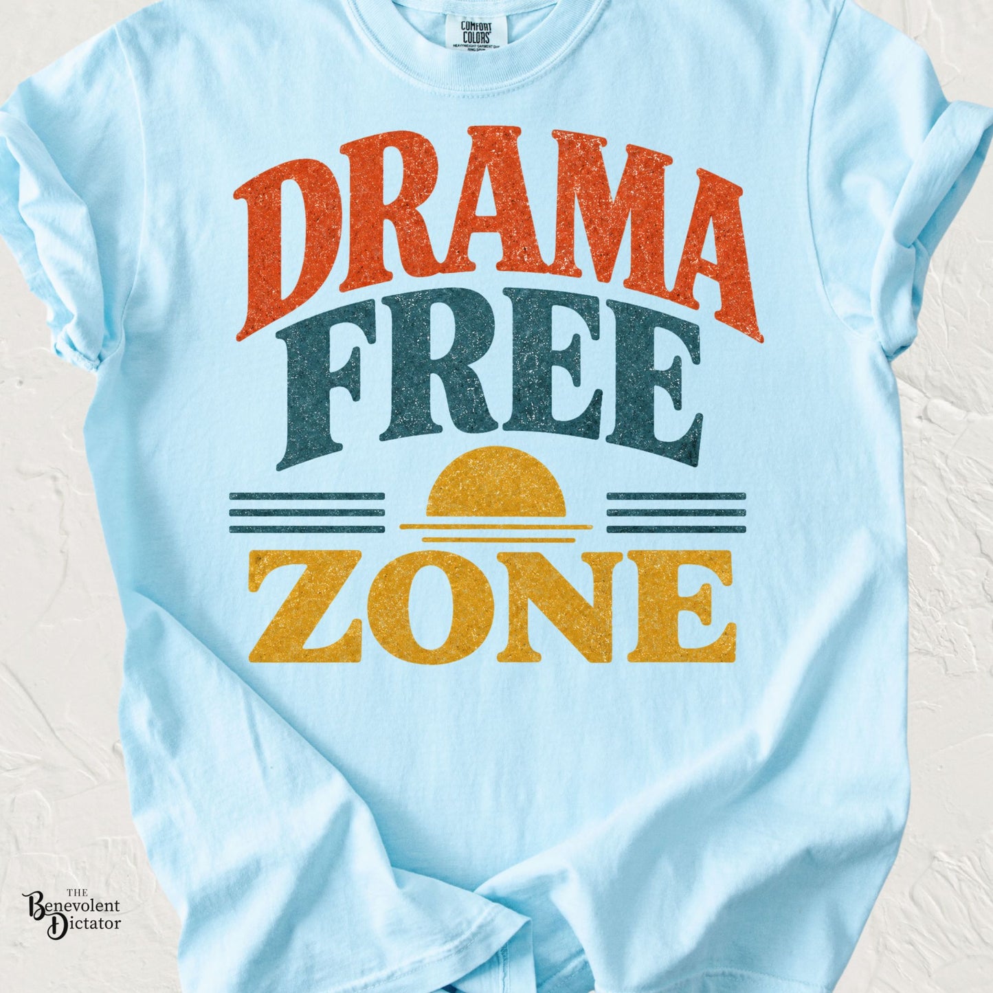 Light blue Comfort Colors t-shirt featuring the phrase "Drama Free Zone" in bold vintage-inspired lettering with a retro sunset graphic in warm orange, teal, and mustard tones.