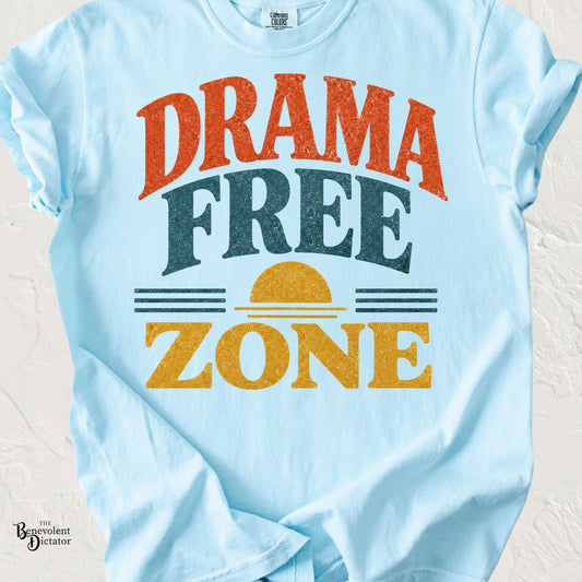Light blue Comfort Colors t-shirt featuring the phrase "Drama Free Zone" in bold vintage-inspired lettering with a retro sunset graphic in warm orange, teal, and mustard tones.