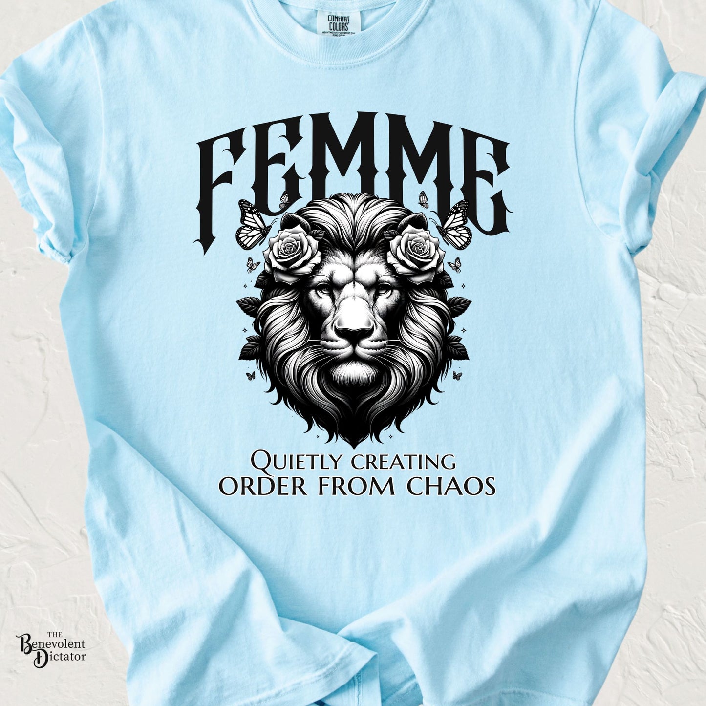 Black-and-white illustration of a regal lion wearing floral roses and surrounded by butterflies beneath the word "FEMME" with the phrase "Quietly Creating Order From Chaos" on a blue Comfort Colors T-shirt.