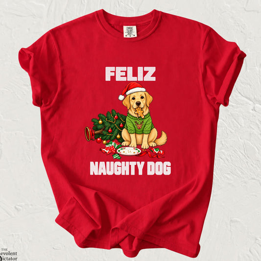 Bright Christmas red Comfort Colors t-shirt featuring a golden yellow Labrador puppy wearing a Santa hat and green reindeer sweater, holding a gingerbread cookie in its mouth. A toppled Christmas tree, scattered ornaments and an empty cookie plate surround the puppy, with festive white lettering reading “Feliz Naughty Dog.”
