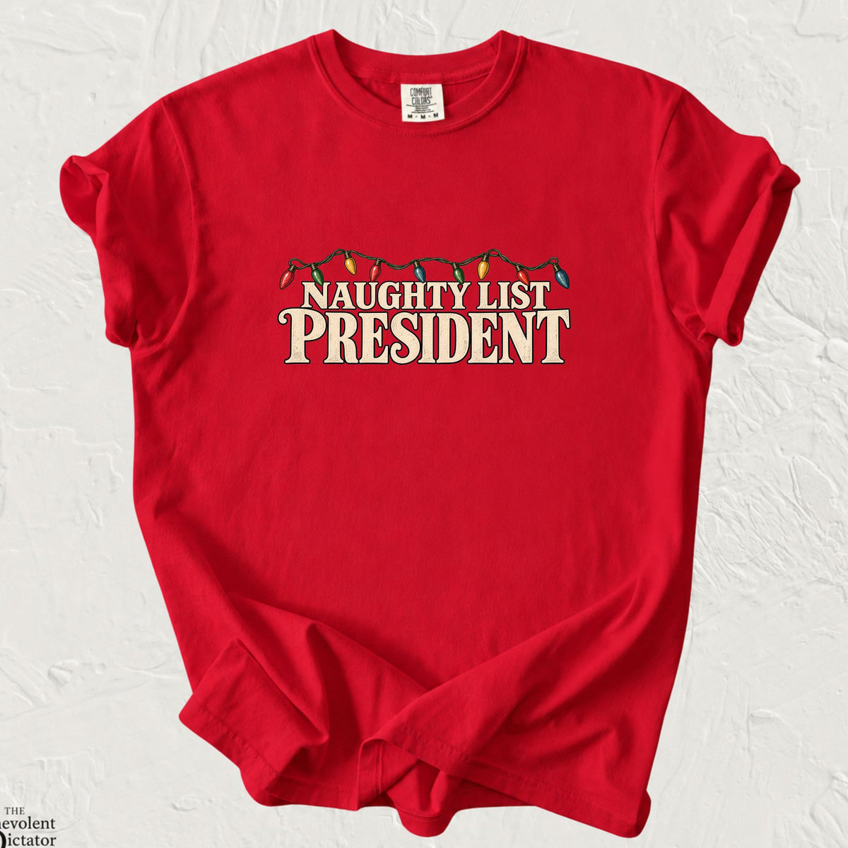 Christmas red Comfort Colors t-shirt featuring vintage-style cream typography reading “Naughty List President,” outlined in black, with a string of colorful red, green, yellow and blue Christmas lights draped above the lettering.