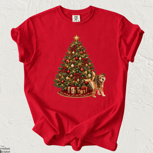 Red Comfort Colors t-shirt featuring a beautifully decorated Christmas tree with glowing warm lights, red and gold ornaments, festive bows, wrapped presents and a golden star. A mischievous scruffy brown dog stands beside the tree with one hind leg raised, marking the Christmas tree.
