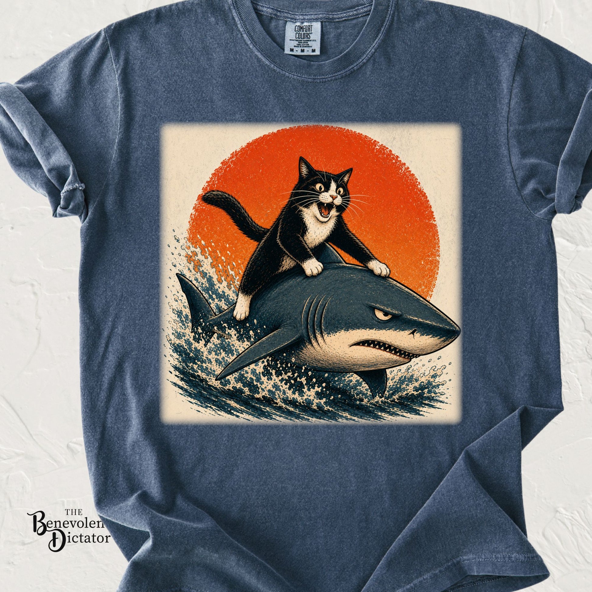 Funny graphic t-shirt featuring a tuxedo cat riding a shark through ocean waves at sunset.