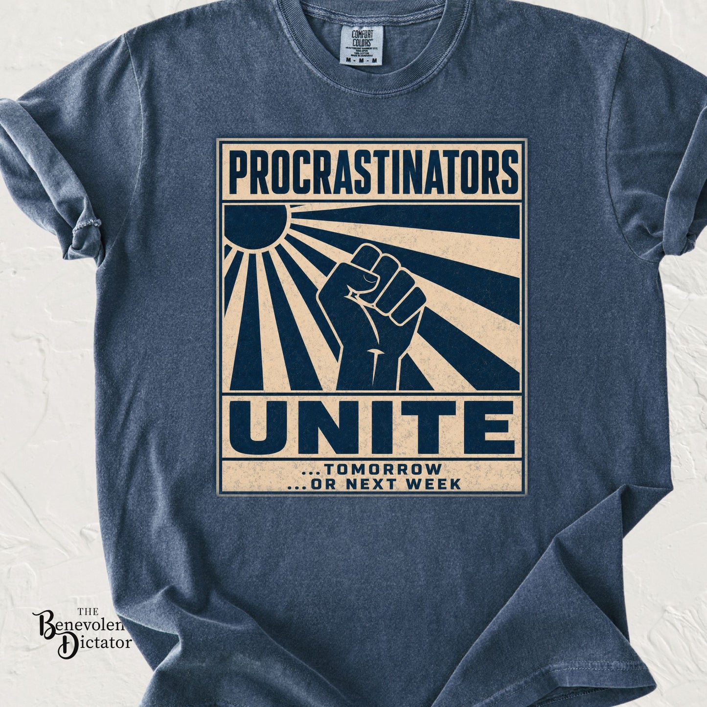 Blue Jean Comfort Colors t-shirt featuring a distressed retro-style graphic with bold cream and navy typography reading “Procrastinators Unite… Tomorrow or Next Week” with a raised fist and sunburst design.